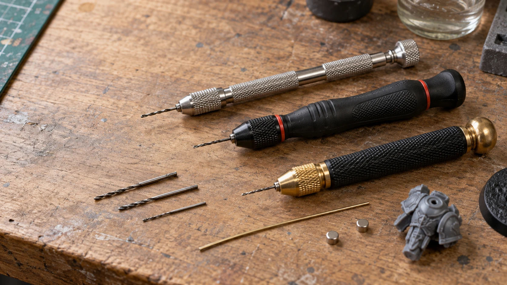 Three silver, black-and-red and black-and-brass manual pin vises with fine drill bits, pinning wire, magnets and a grey miniature part
